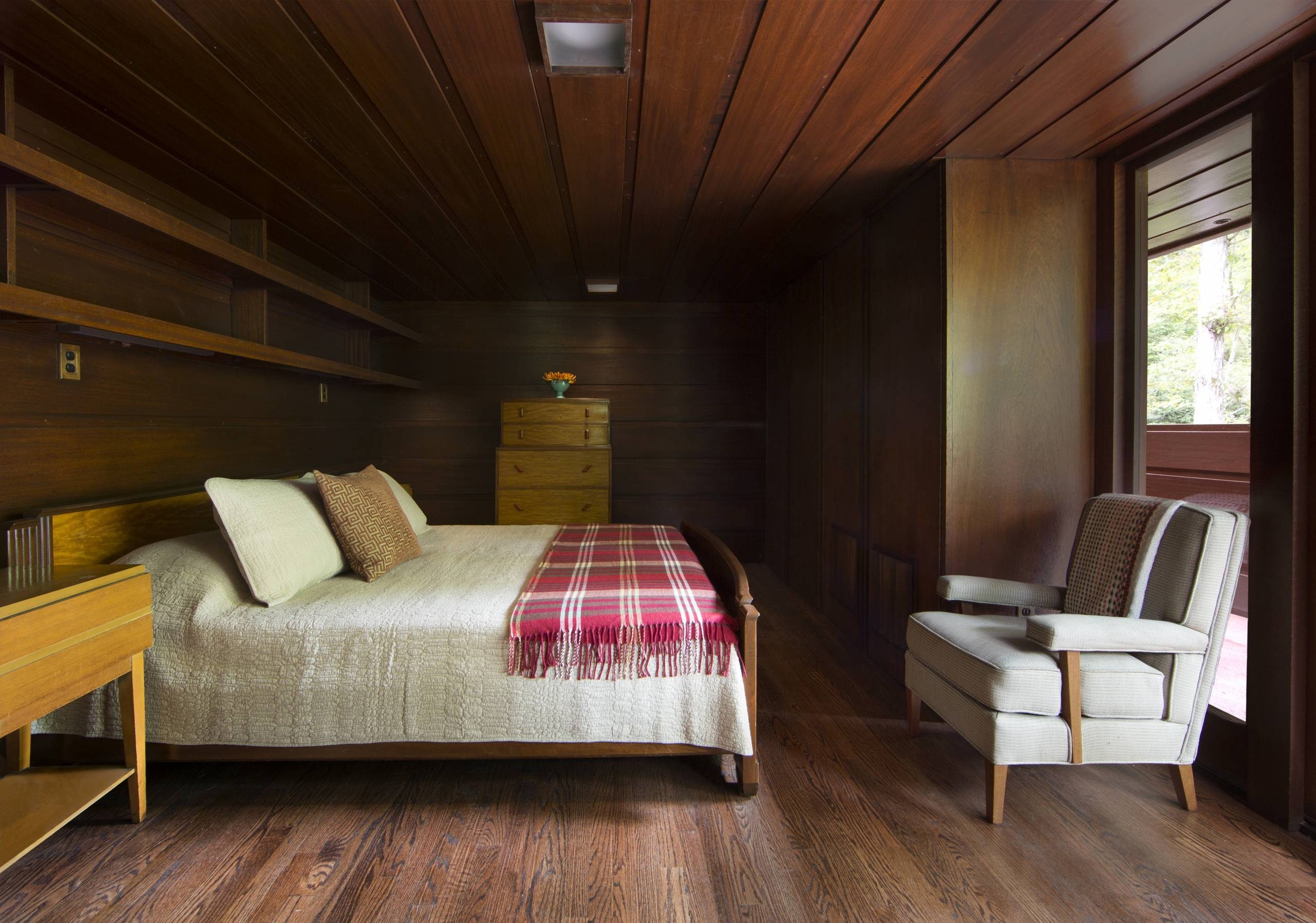 Frank Lloyd Wright Bachman-Wilson House Master Bedroom Bedroom with wooden paneling, double bed, quilt, plaid blanket, armchair, drawers, shelves.