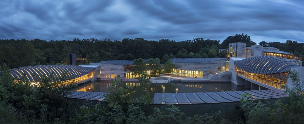 Modern architectural complex with curved, glass structures surrounded by water and trees at dusk.