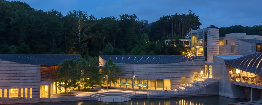 Crystal Bridges at Dusk Modern architectural complex with curved roofs, large windows, forest, and water reflections at dusk.