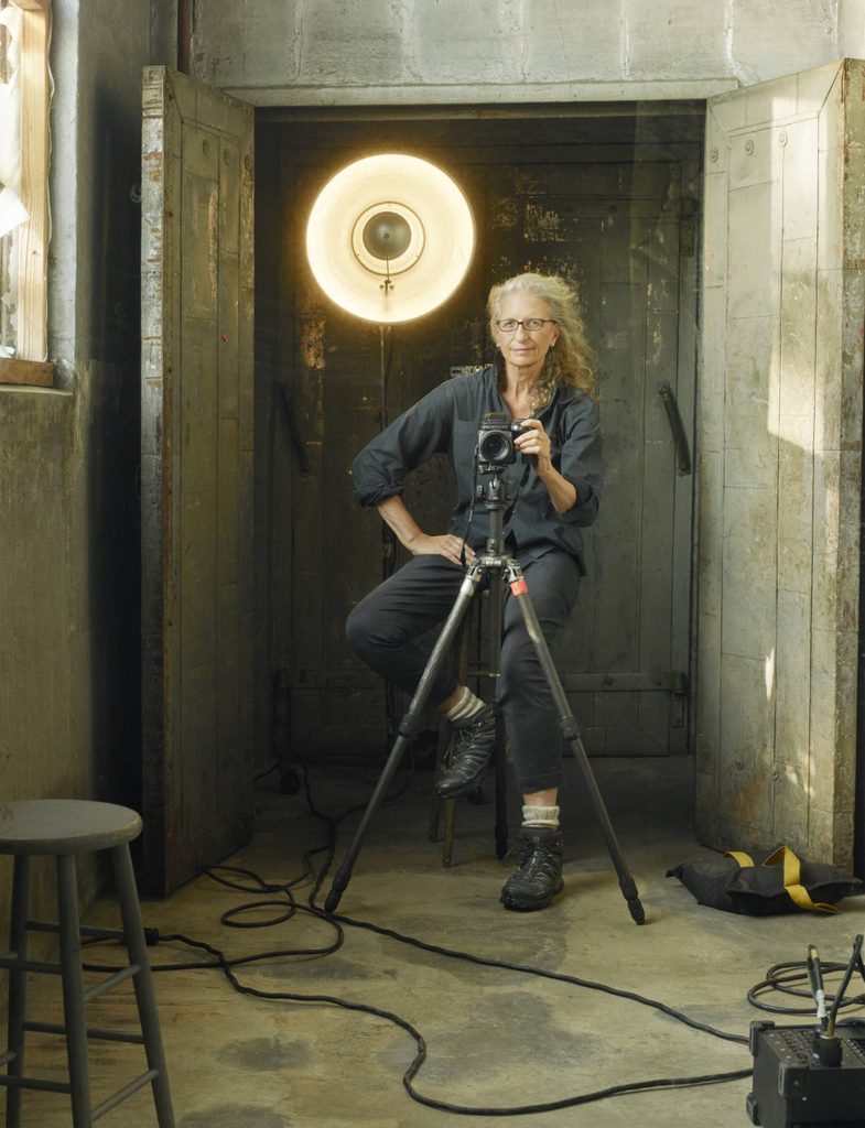 Annie Leibovitz, Self Portrait, Brooklyn, New York, 2017. Photo courtesy of the artist ©Annie Leibovitz. Women sits on stool in doorway with larger light behind her and camera on tripod in front of her.