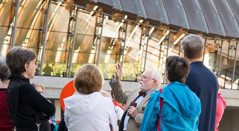 arch_tour Tour guide gesturing to an attentive group outside a reflective modern building.