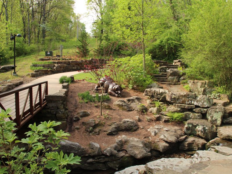 Art Trail Serene garden with a wooden bridge, rocky landscape, lush greenery, and stone pathways.