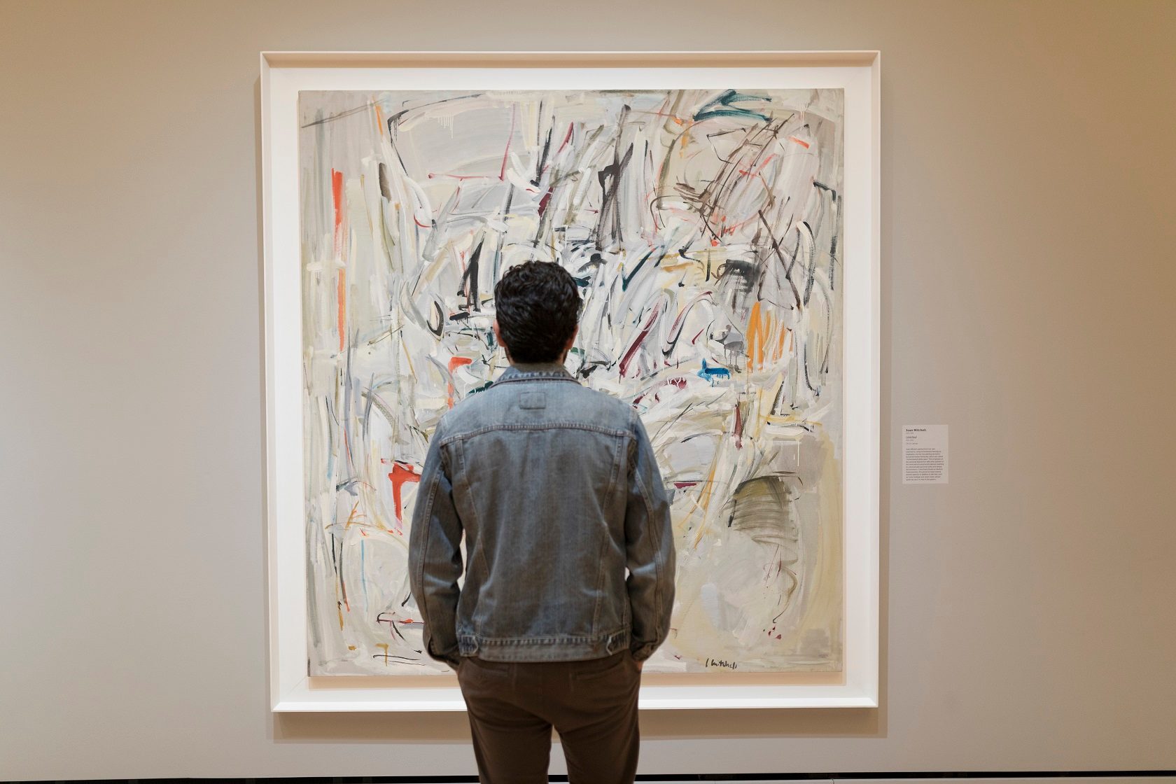 Man looks at abstract painting Person in denim jacket views colorful abstract painting in gallery with info plaque.