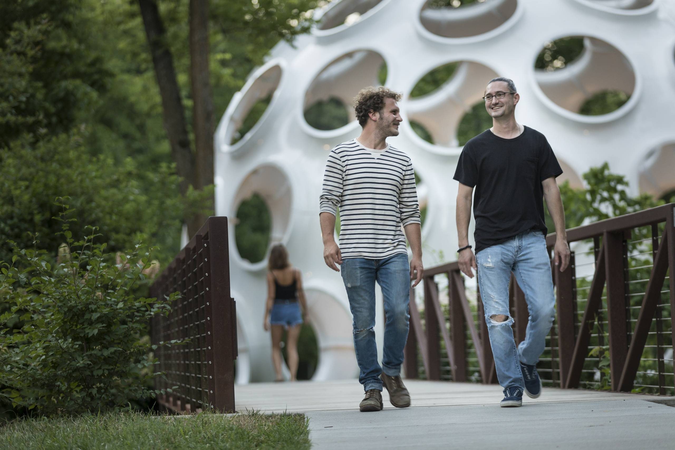 Guests walking on Trail Two people walking on a bridge near greenery and a circular architectural structure.