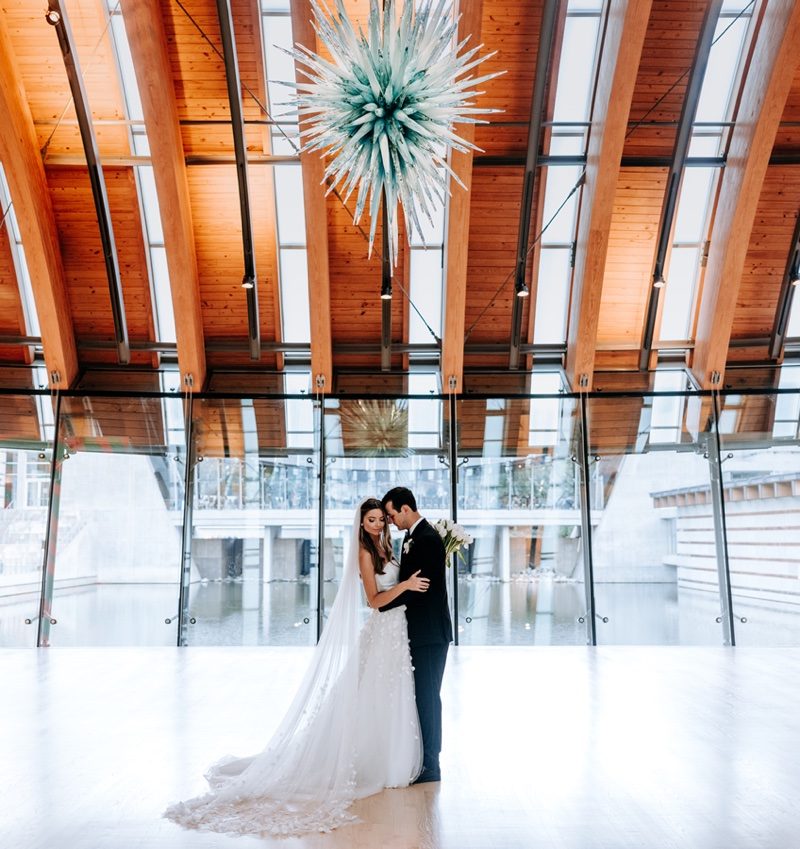 Wedding Venue Bride and groom embrace in modern space with glass walls, wooden beams, and spiky chandelier.