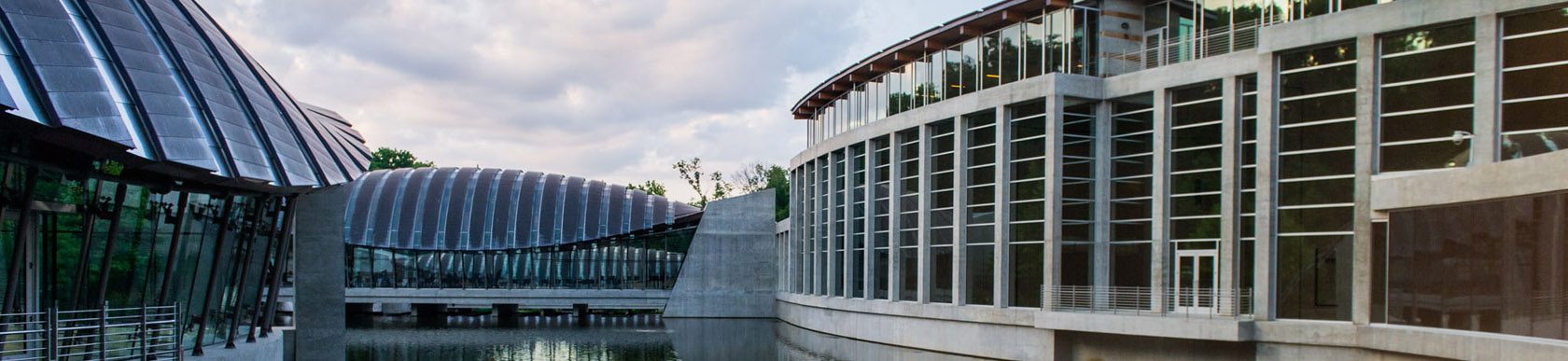View of Crystal Bridges and water feature Modern architectural complex with curved glass, large windows, water reflection, and greenery.