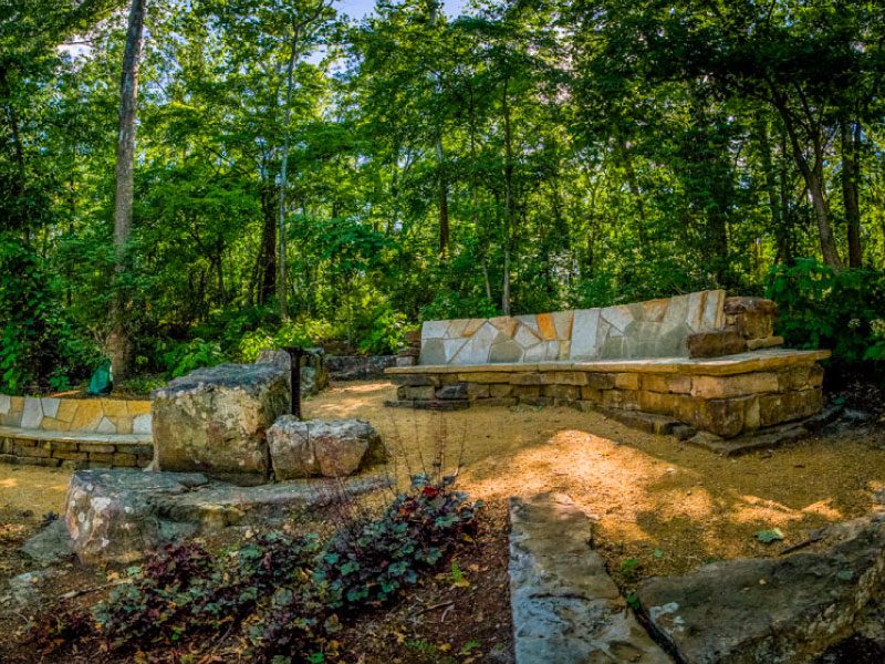 Crystal Bridges Overlook Outdoor stone seating area surrounded by trees and foliage, with a stone wall and dirt clearing.