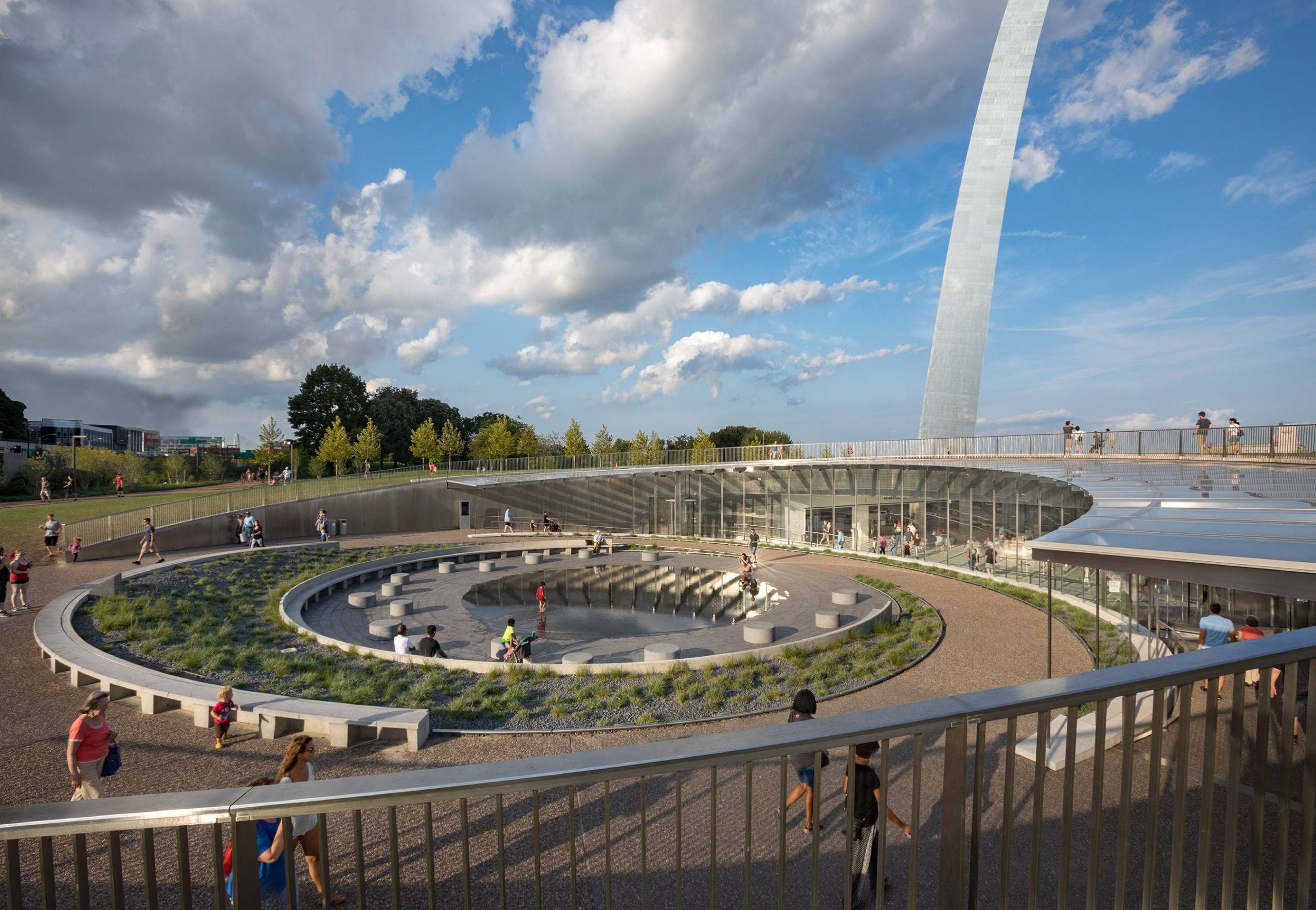 Circular Park with Reflective Pool Modern outdoor space with circular seating, landscaping, people, pool, and upward arch structure.
