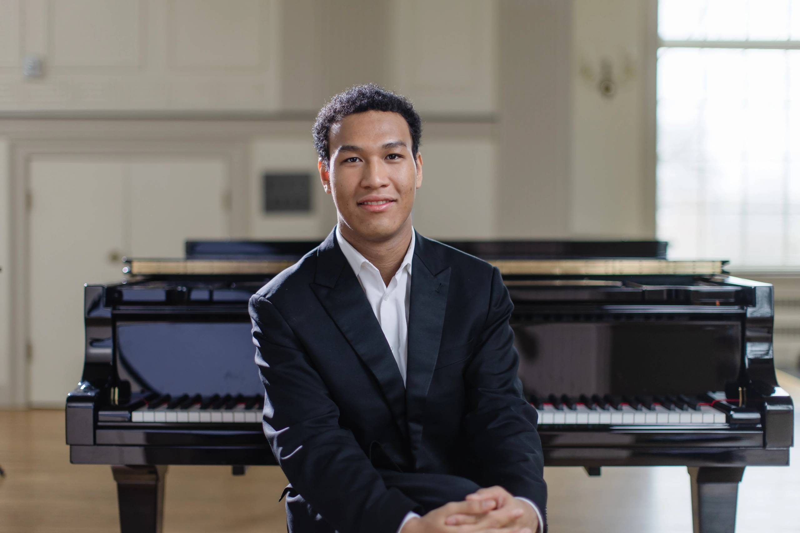 Pianist in Formal Setting Person in a suit seated by a grand piano in a bright room.