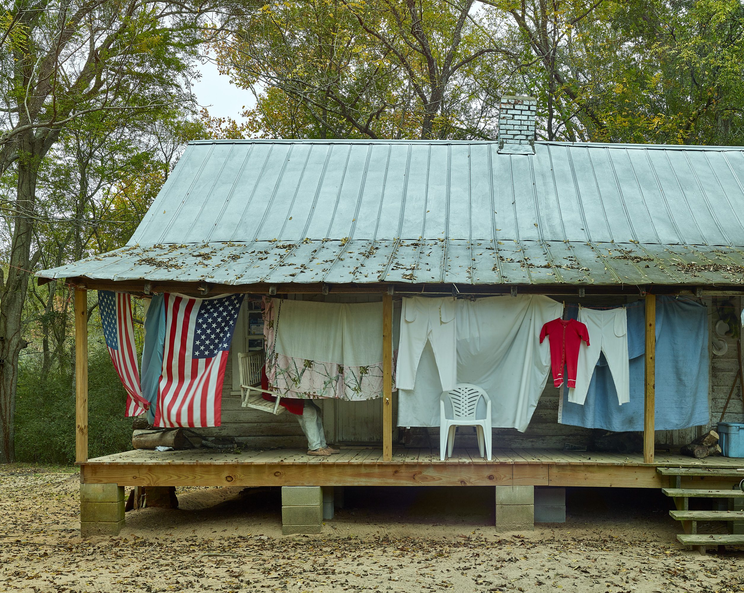 Cabin with Laundry and Flags Rustic cabin with tin roof, porch, laundry, flags, and a white chair amid trees and fallen leaves.