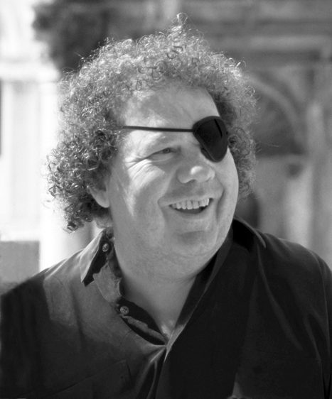 Dale Chihuly Curly-haired person with eyepatch and dark shirt smiling outdoors, urban background blurred.