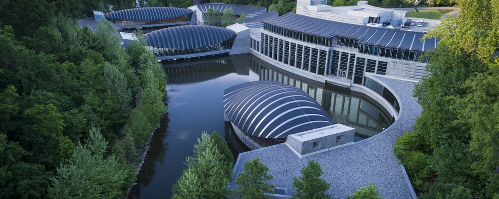 Overhead view of Crystal Bridges Museum of American Art Modern architectural complex with curved roofs amid greenery, encircling a reflective water body.