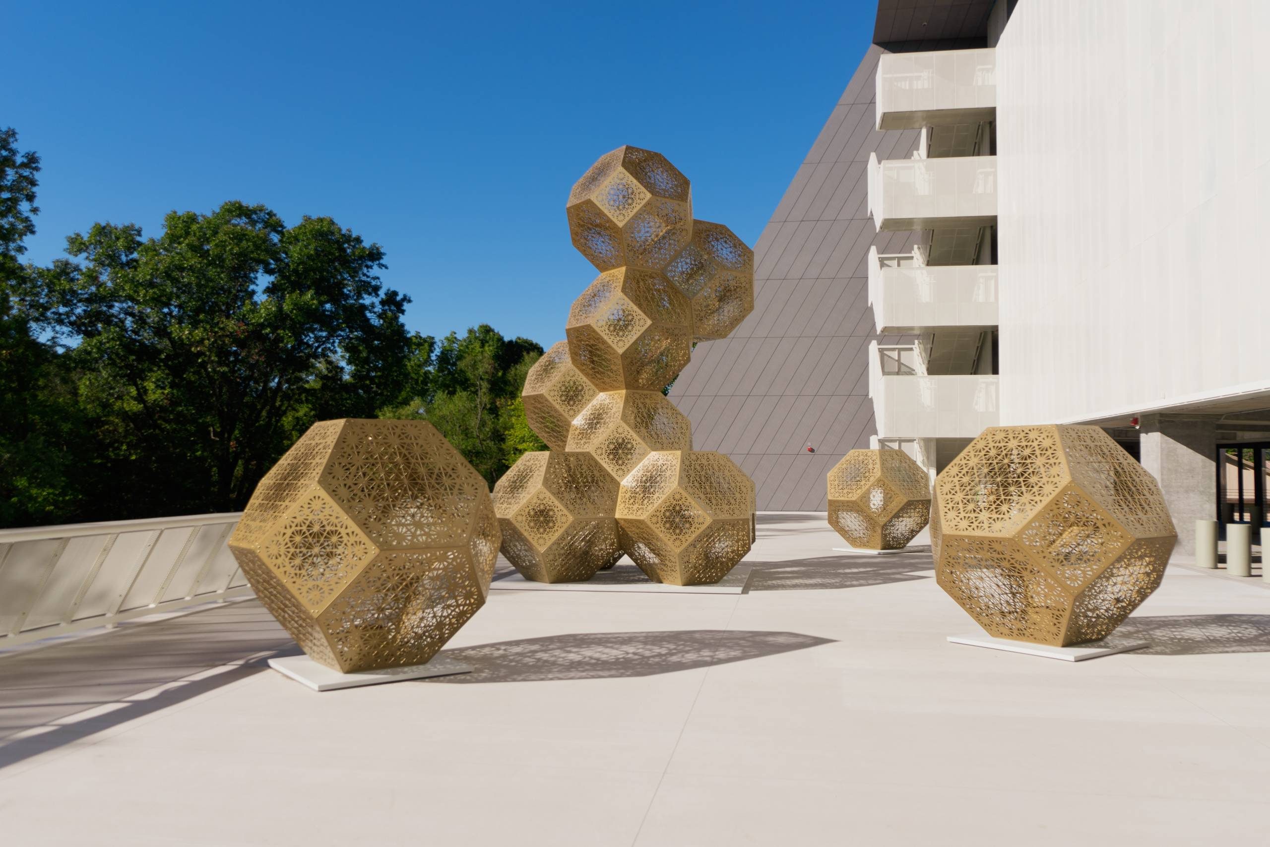 HYBYCOZO, Congruence, 2023, powder coated stainless steel and LED lighting, large cluster: 21 ft. x 12 ft. x 12 ft.; spheres: 6 ft. x 6 ft. x 6 ft. Courtesy of the artists. Outdoor installation with geometric metal sculptures on concrete near a modern building.