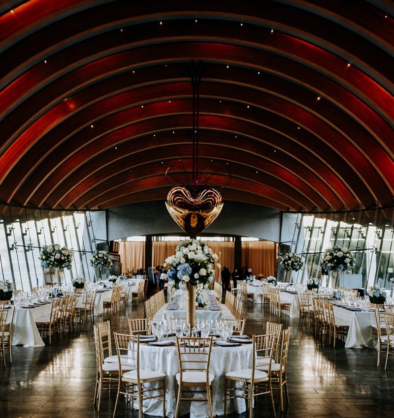 Reception Reception hall with round tables, white tablecloths, gold chairs, floral centerpieces, large windows.