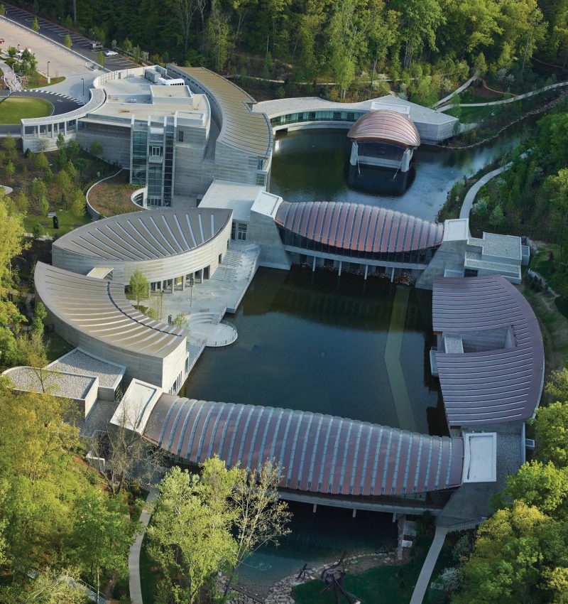 Crystal Bridges aerial view Aerial view of curved architectural complex around a pond, surrounded by greenery and walkways.