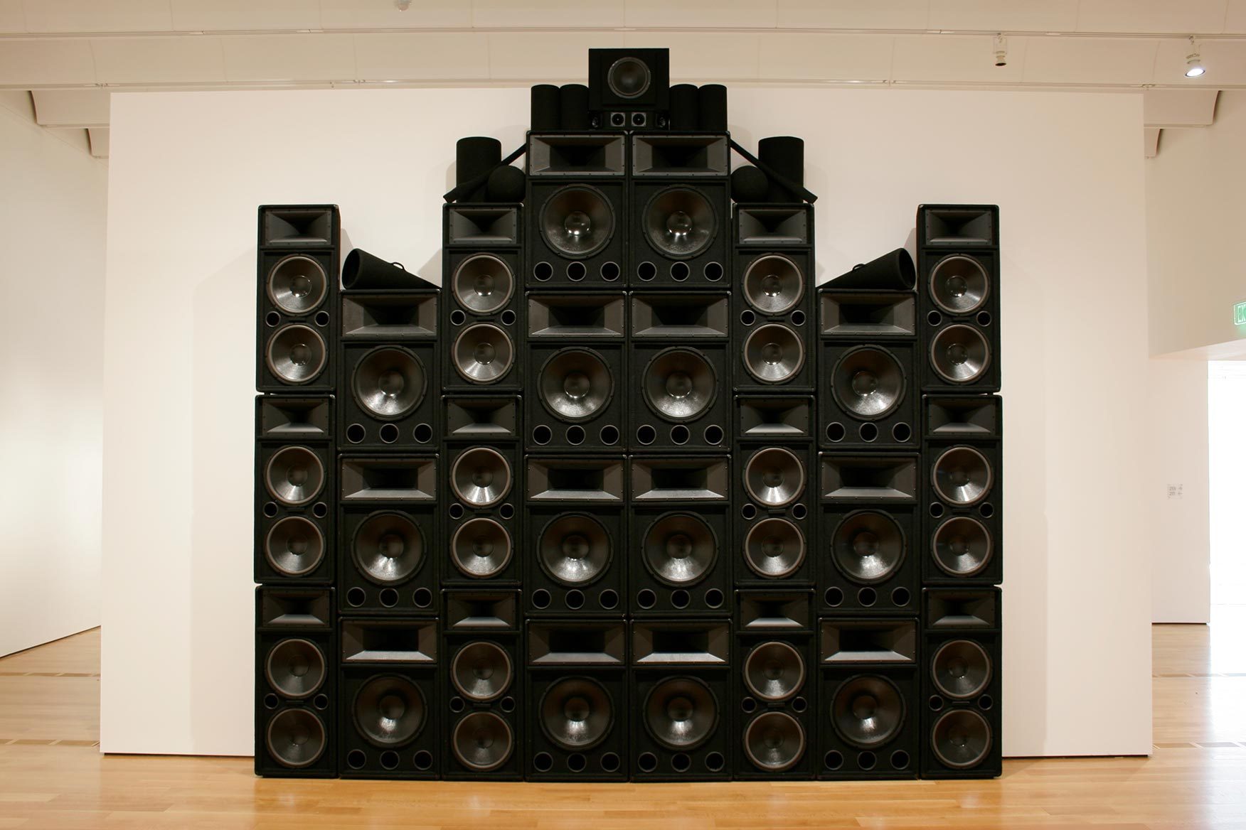 Nadine Robinson, Coronation Theme: Organon, 2008, speakers, sound system, and mixed media, 175 x 18 1/2 x 174 in. High Museum of Art, Atlanta, given by John F. Wieland, Jr. in memory of Marion Hill, 2008.175 A large structure of stacked black speakers, black megaphones and black cylinders towers next to a white wall