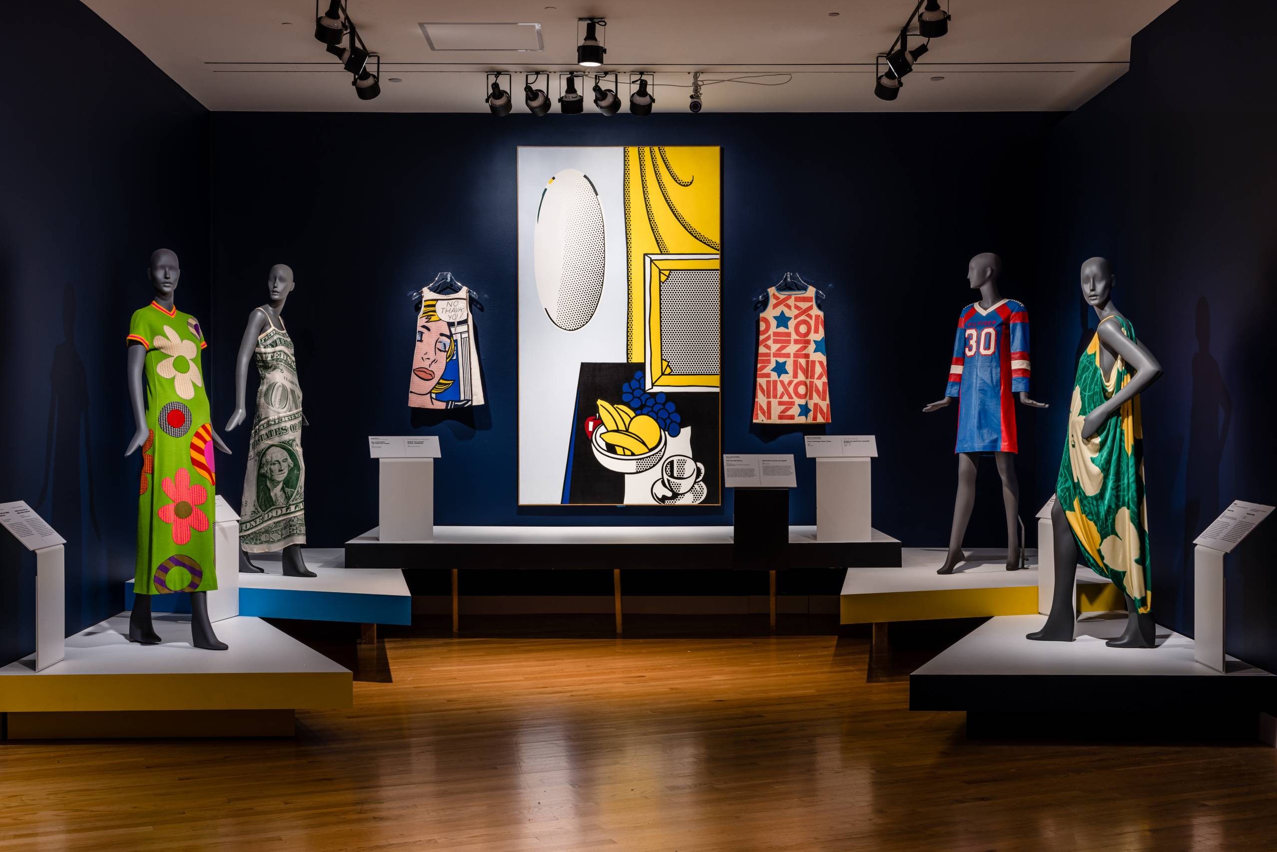 Fashioning_America-327 (1) a gallery space featuring mannequins dressed in pop art-inspired garments with bright colors and patterns framing Roy Lichtenstein's work