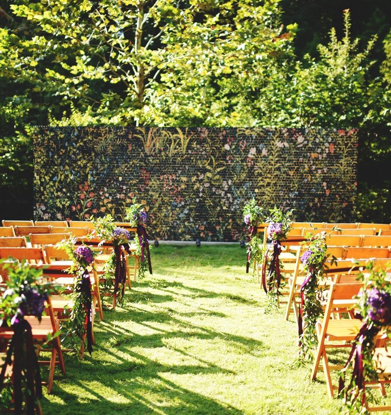 Wedding Venue Outdoor event setup with decorated chairs, floral wall, and trees in the background.