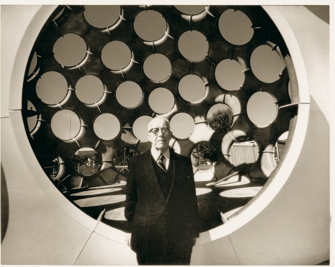 R. Buckminster Fuller (1895 - 1983), Fly's Eye Dome, 1961, fabricated ca. 1980, Fiberglass-reinforced polyester, 38 × 50 × 50ft. Crystal Bridges Museum of American Art, Bentonville, Arkansas, 2015.15 Elderly man in formal attire before a structure with circular openings in sepia tones.