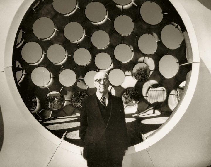 R. Buckminster Fuller (1895 - 1983), Fly's Eye Dome, 1961, fabricated ca. 1980, Fiberglass-reinforced polyester, 38 × 50 × 50ft. Crystal Bridges Museum of American Art, Bentonville, Arkansas, 2015.15 Older man in suit stands before circular structure with circles, trees and fence in background.
