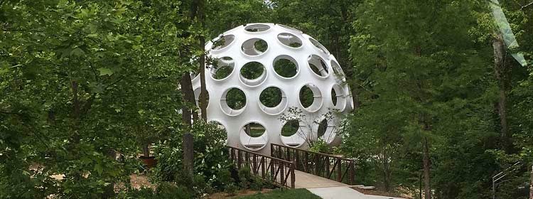 R. Buckminster Fuller (1895 - 1983), Fly's Eye Dome, 1961, fabricated ca. 1980, Fiberglass-reinforced polyester, 38 × 50 × 50ft. Crystal Bridges Museum of American Art, Bentonville, Arkansas, 2015.15 Spherical structure with circular cutouts in a forest, accessed via a wooden bridge.