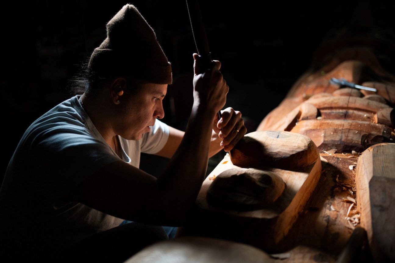 Nicholas Galanin Person carving a wooden sculpture with a chisel in dim light, surrounded by wood shavings.