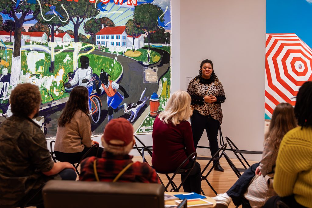 Person presenting to seated group by mural of scenic street with children playing indoors.