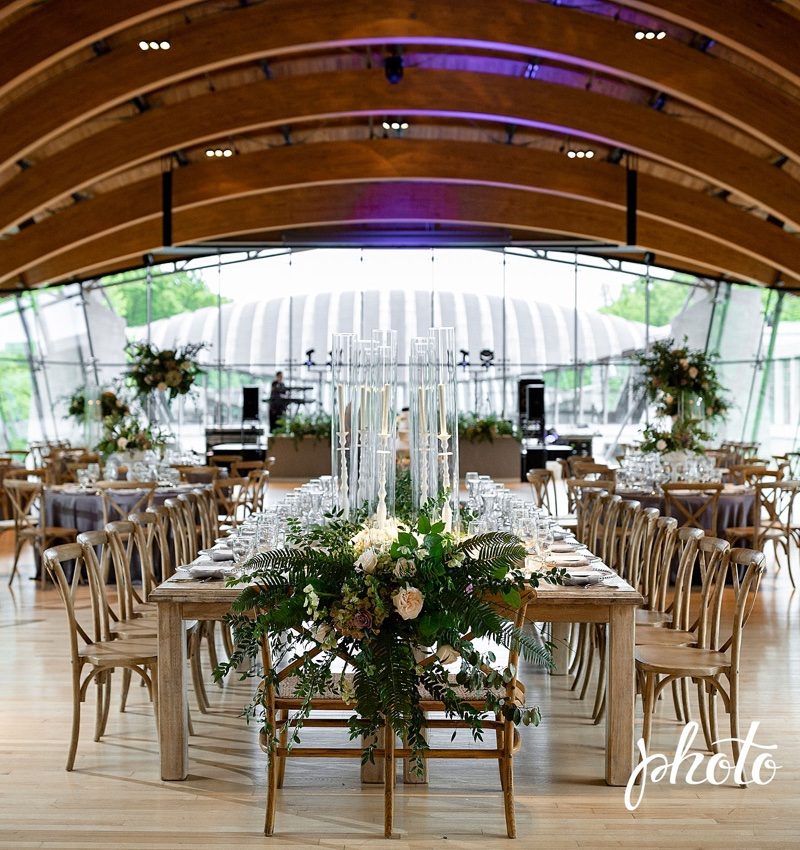 Great Hall Reception Elegant dining venue with wooden tables, floral decor, glass candles, arched ceiling, large windows.