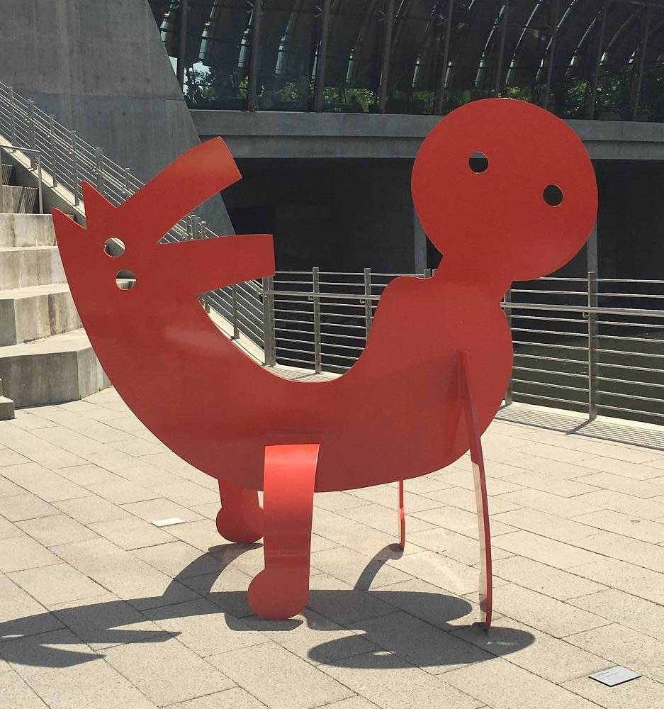 Keith Haring, Two-Headed Figure, 1986, polyurethane paint on aluminum. Crystal Bridges Museum of American Art, Bentonville, Arkansas Red abstract metal sculpture of reclining figure with round head and circular eyes on paved area.