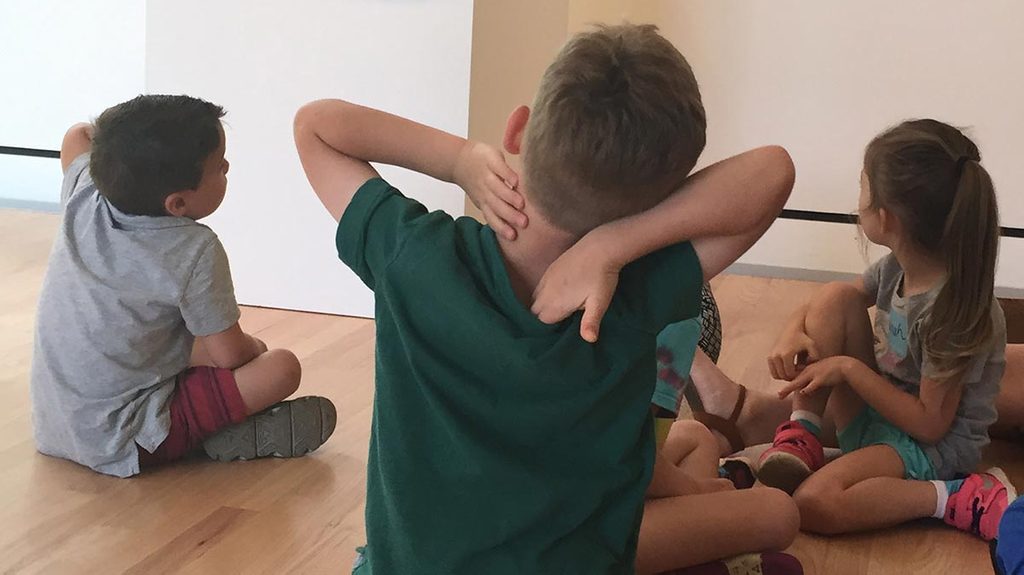 header-community-youth-tours-crop Children sitting on a wooden floor in an informal setting, one with hands on head.
