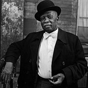 headshot-bey-dawoud Older man in bowler hat and suit with bow tie standing by brick wall and railing.