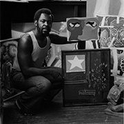 headshot-chandler-dana Man in studio with paintings, including abstract art with a star and facial features, sitting on floor.