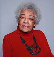 headshot-jarrell-jae Elderly woman with gray hair, red turtleneck, black necklace, plain background.