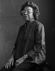 headshot-lawrence-carolyn Elderly woman seated, wearing glasses and patterned top, in a monochromatic setting.