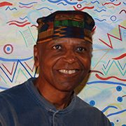 headshot-williams-gerald Person in patterned hat and dark shirt smiling in front of colorful abstract background.