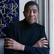 headshot-williams-t-william Person in black turtleneck with crossed arms, standing beside abstract artwork on white wall.