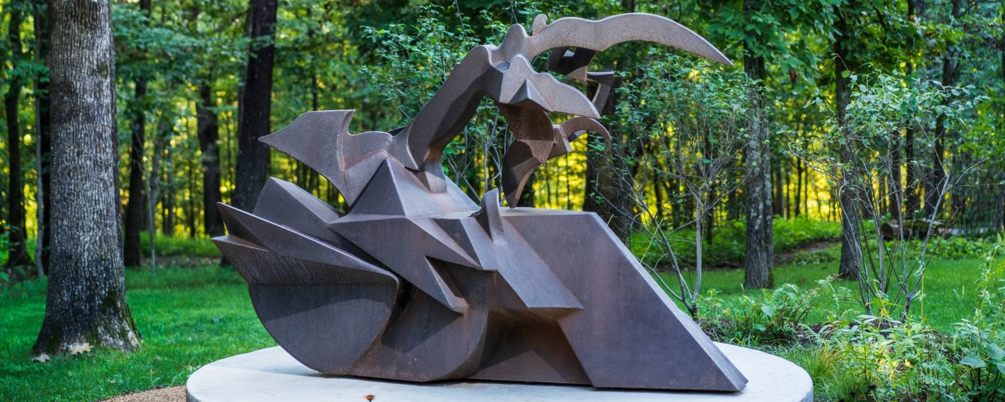 Hunt_SlowlyTowardsTheNorth-1_6000px-scaled_1990x796_acf_cropped-1 a steel gray sculpture shaped like a train engine and wheel on the bottom with a scythe shape on top sitting on a platform surrounded by a lush green forest
