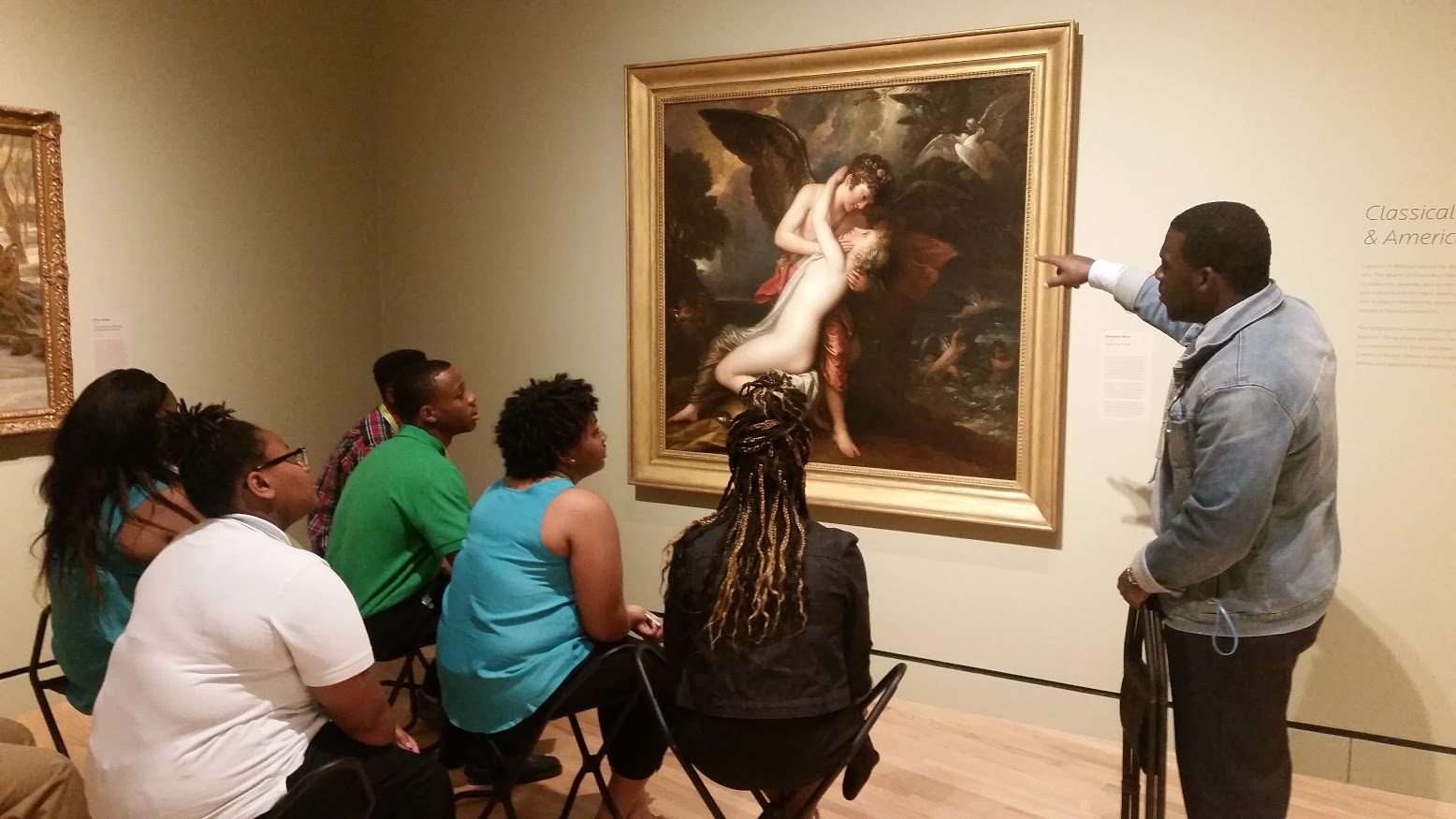 Intern discuss an artwork in the galleries Group in museum listens to guide explaining a classical painting, pointing at details.