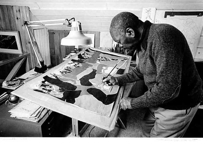 Jacob Lawrence Man painting details on a large canvas in a studio with desk lamp and art materials.