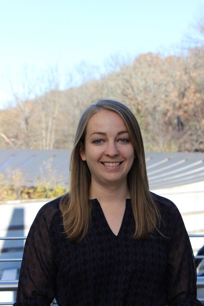 Jessi Mueller Person with long hair and dark blouse smiling outdoors, bare trees and clear sky in background.
