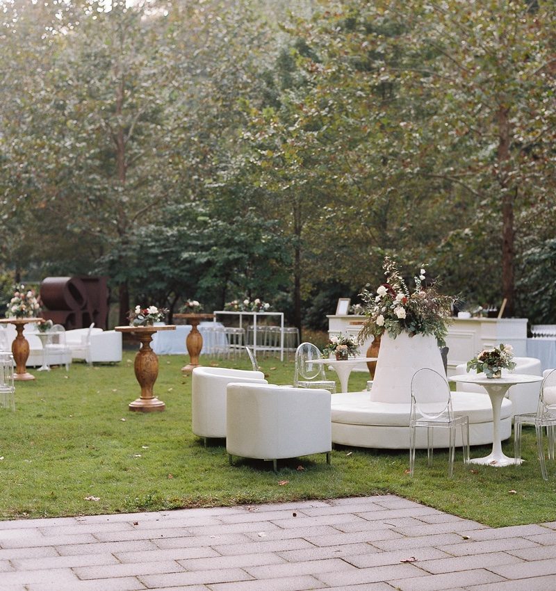 Reception Outdoor event with white furniture, floral arrangements, on grass surrounded by trees.