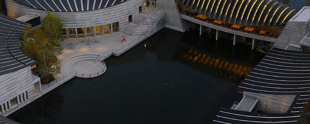 Aerial View of Crystal Bridges Modern curved buildings with dark roofs, glass windows, wooded autumn area, large water body.