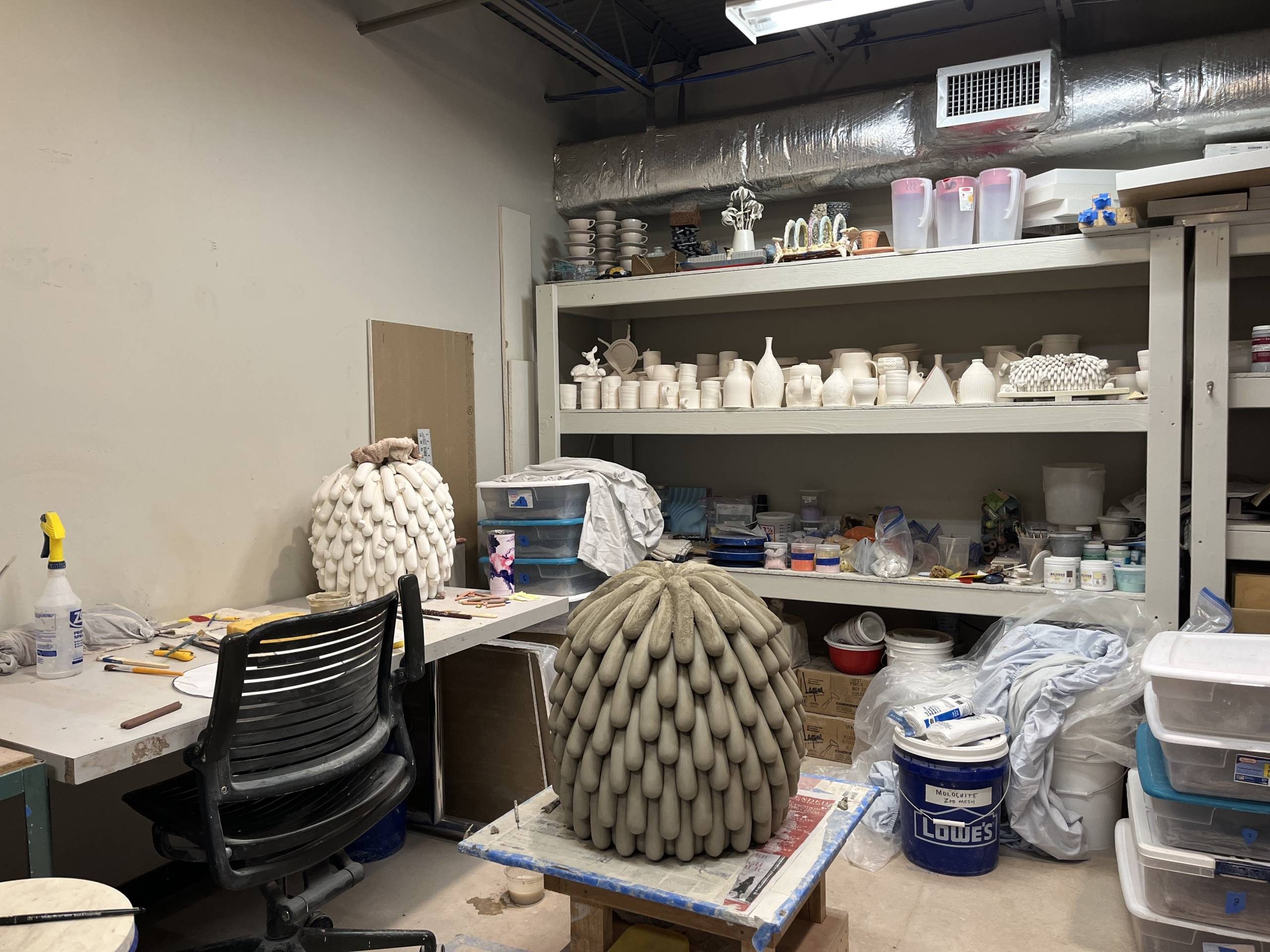 linda3 an artist studio with a gray clay work in progress on a workbench in the center, a work desk to the left, and shelves filled with white clay jars and art supplies in the background