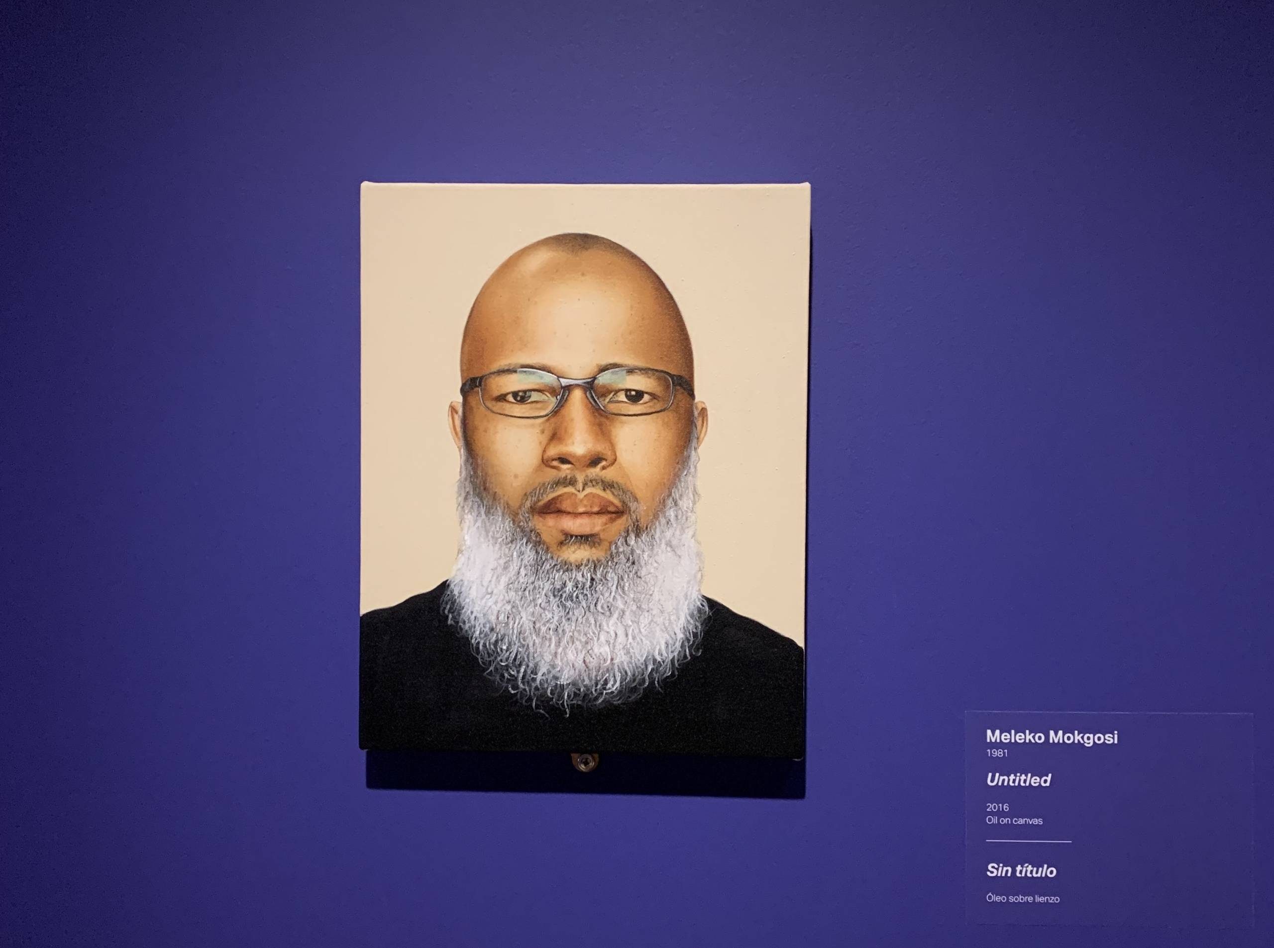 Meleko Mokgosi, Untitled, 2016, oil on canvas, 14 × 11 × 1 in. Crystal Bridges Museum of American Art, Bentonville, Arkansas, 2020.15. Self-portrait of man with grey beard wearing glasses hanging on wall with exhibition labels beside it in Crystal Bridges at 10