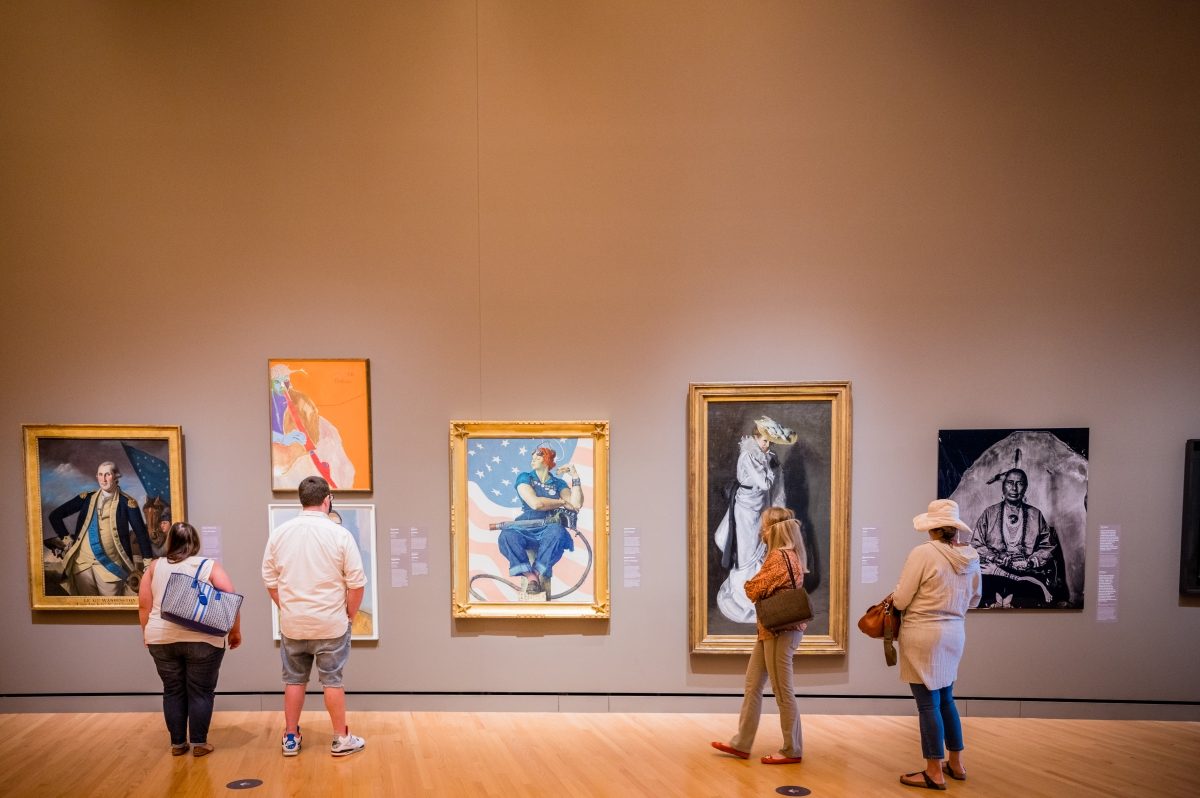 Guests in the Early American Art Gallery People viewing various framed artworks in a gallery with wooden floors and neutral walls.
