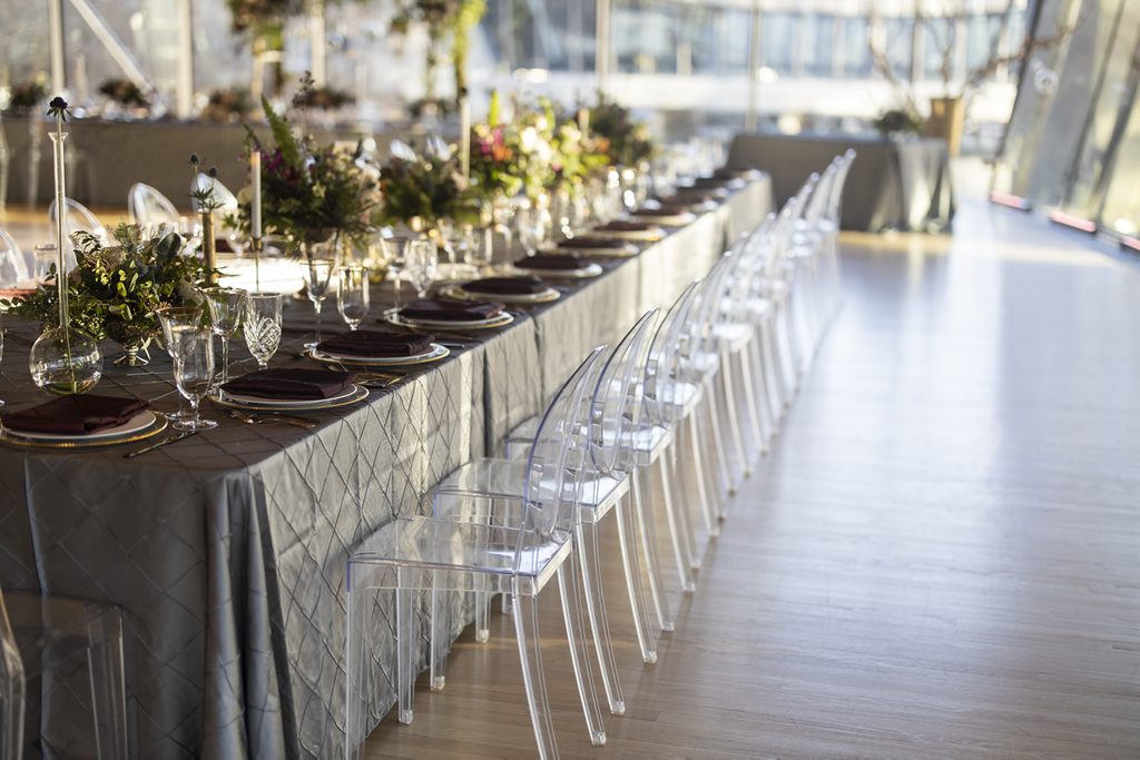 Reception Elegant dining table with floral centerpieces, quilted tablecloth, and transparent chairs.