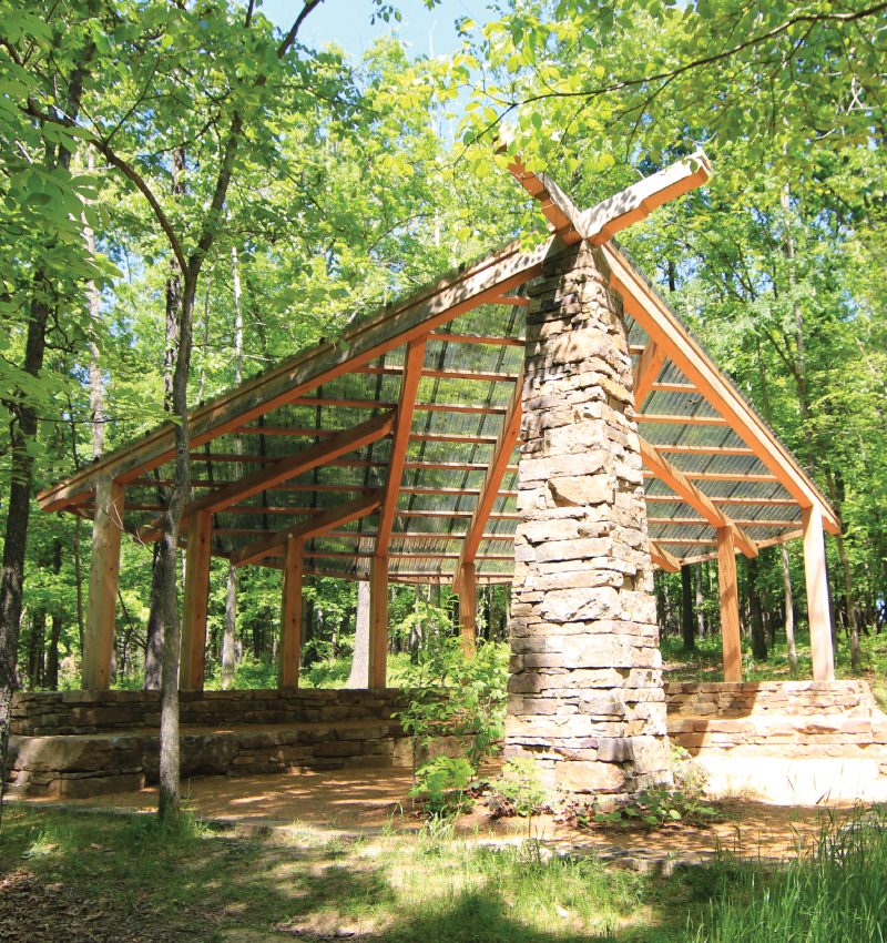 North Forest Shelter Wooden pavilion with stone pillar and transparent sloped roof in a lush forest setting.