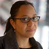 Sandra-Jackson-Dumont.jg_ Person with glasses in dark coat looks at camera; blurred background.