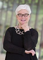 Sandy-Edwards-sm Individual with white hair, glasses, smiling in black outfit and necklace; hand near chin.