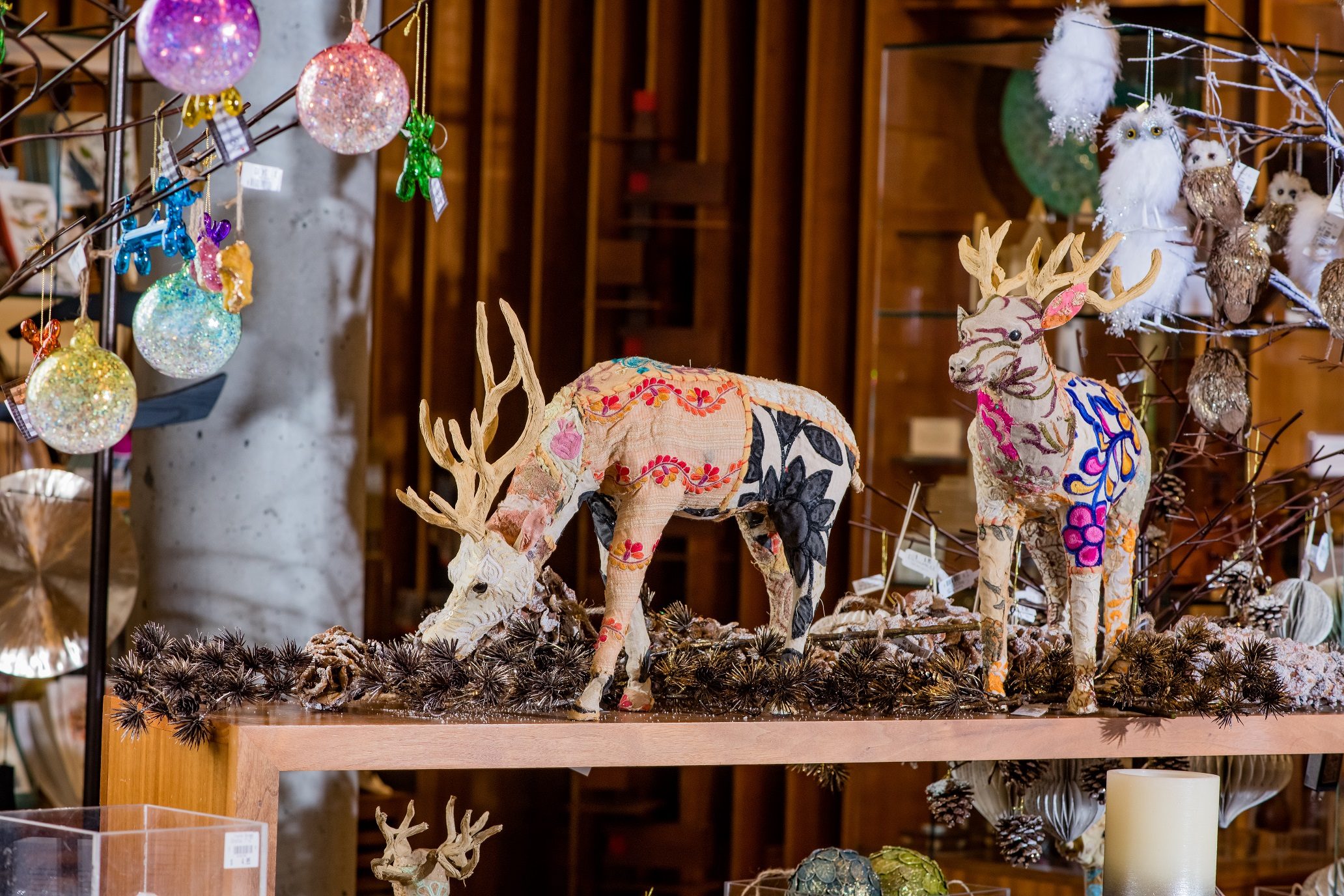 Merchandise Decorative deer figures with colorful patterns on a shelf, surrounded by ornaments and pinecones.